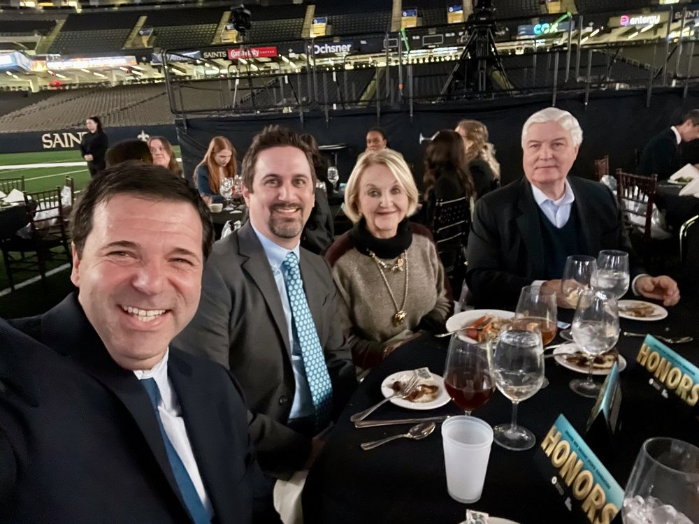 Reid Stone, Shaun Walker, Diane Lyons, and her husband Jack Lyons seated at a table during the Greater New Orleans Sports Foundation Honors Luncheon inside the Superdome. Plates, drinks, and “HONORS” table signs are visible, with the Saints field in the background.