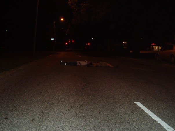 Two friends lying in the middle of a dark, empty street in Lakeview, New Orleans, after Hurricane Katrina. The street is quiet with no lights or visible activity, reflecting the stillness of the city.