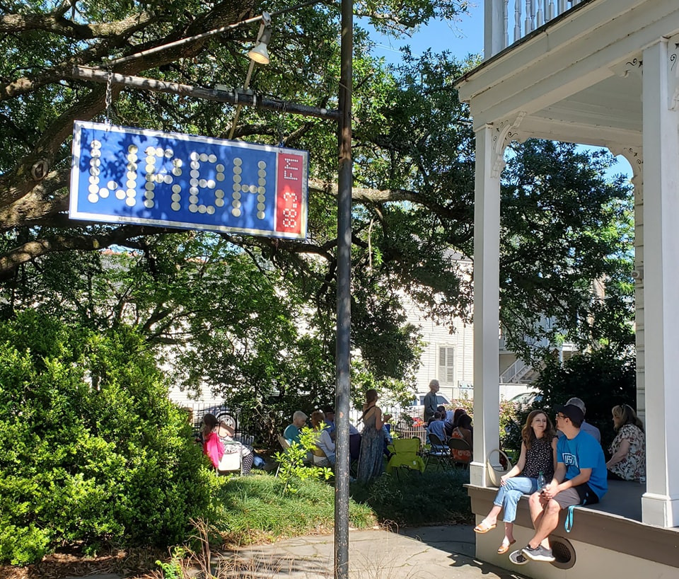 People sitting on the porch of the WRBH Reading Radio studio on Magazine Street in New Orleans with the WRBH logo sign shown on a warm, sunny spring day.